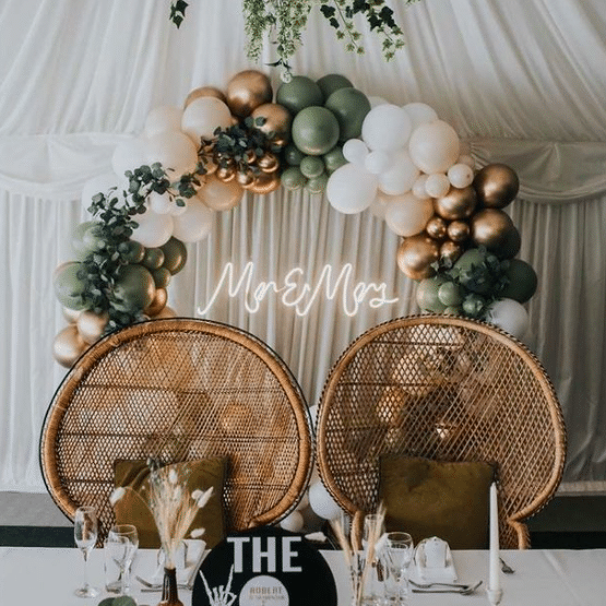 Neon wedding sign displaying the words 'Mrs & Mrs' written in a cursive font in a warm white lighting. The sign is displayed within a gold and green balloon arch behind a pair of round rattan chairs. 
