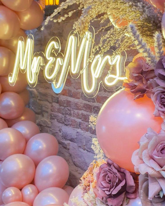 Mr and Mrs warm white neon sign displayed within a pink balloon and flower hoop against a brick wall