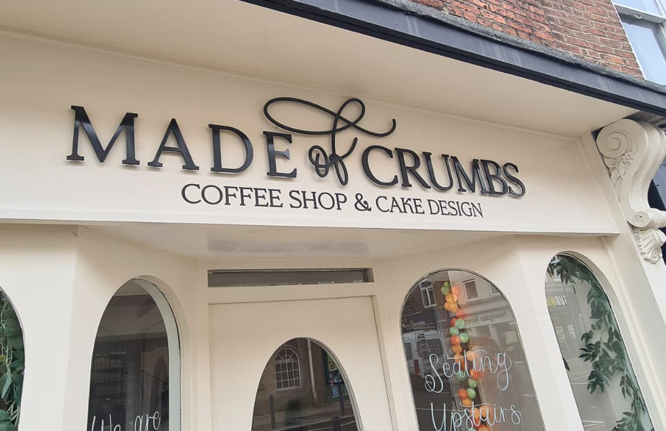 Signage for coffee shops - Image shows the front of a coffee shop based in Morpeth, called Made Of Crumbs. The shop exterior is painted cream, and has 4 large arched windows. The sign features a black logo reading 'Made of Crumbs - Coffee shop and cake design', made from flat cut letters and installed with offset fixings. 