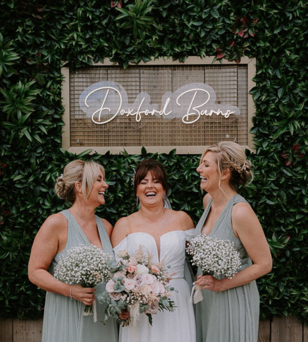 Selfie Wall at Doxford Barns showing a green foliage wall with a caged LED sign reading 'Doxford Barns' in a script font and warm white LED. There are three smiling bridesmaids in front of the selfie wall holding flower bouquets. 