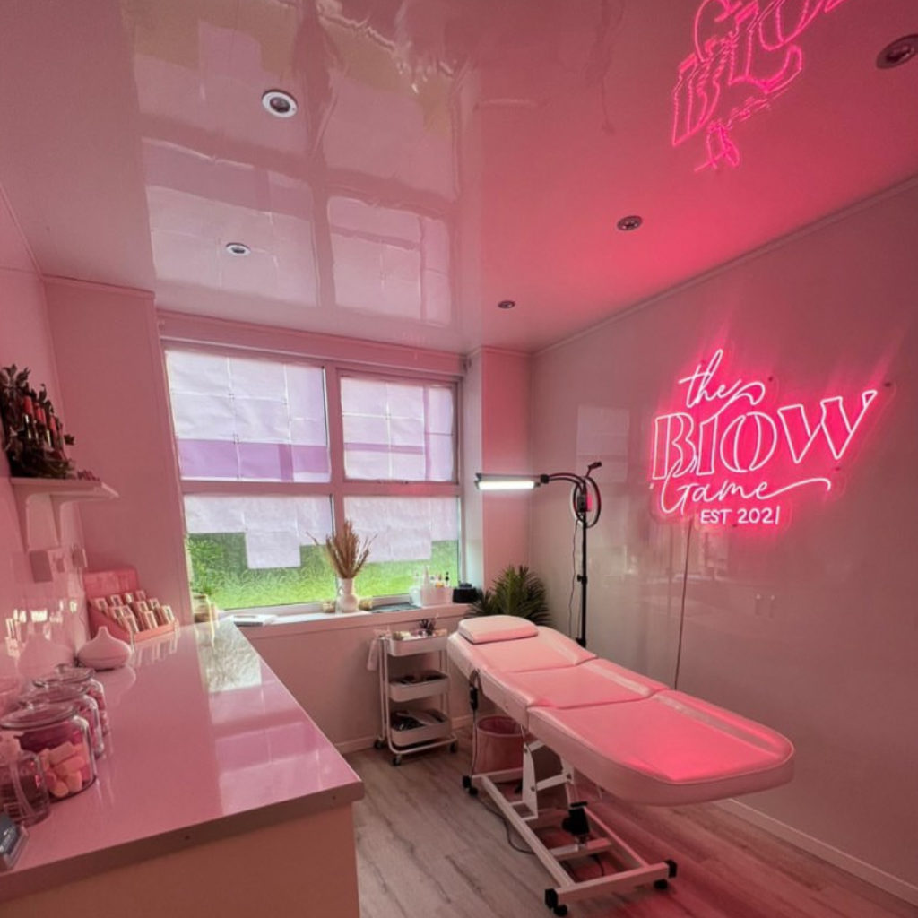 White beauty clinic with a pink LED neon sign on the wall reading 'the brow game' with a pink glow emitting from the sign.