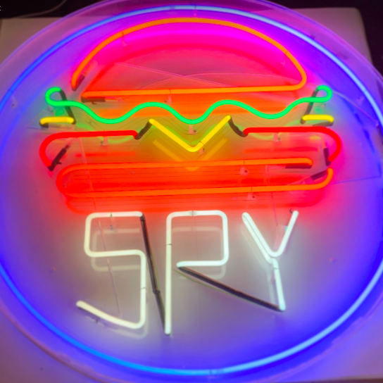 Traditional neon sign for restaurant - neon sign shows an orange burger bun with yellow and red fillings, with the word 'spy' written underneath in white neon. The burger and name have a blue neon ring around them. 