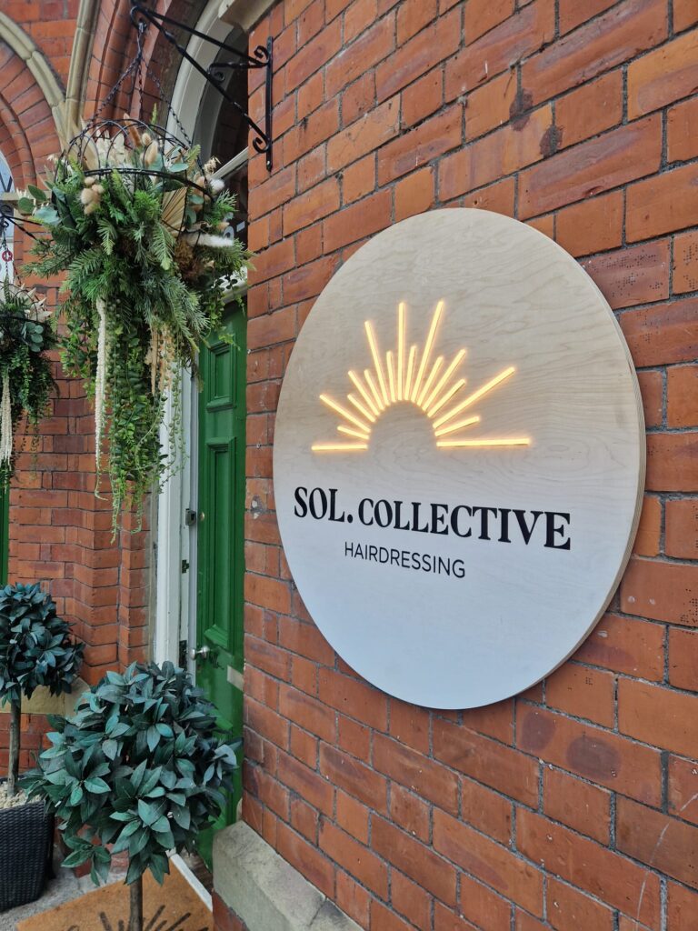 Round wooden sign with a digitally printed white gradient and black acrylic letters reading 'Sol Collective Hairdressing'. Above the black lettering is a warm white LED neon sun logo. The sign is fixed to a red brick wall with brass locators. 