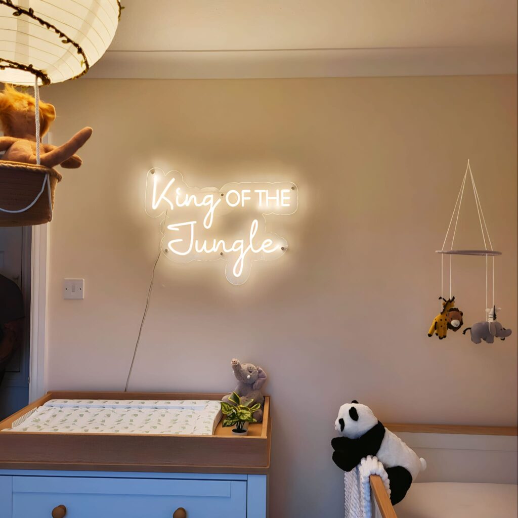 Beige nursery walls with a warm white LED neon sign reading 'king of the jungle' above a wooden baby changing station. 