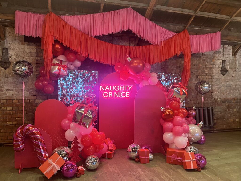 completed event styling setup with pink wooden arches, silver shimmer walls, pink and red tassel garlands hanging from the ceiling, pink and red balloon clusters, disco balls laid on the floor in front of arches 