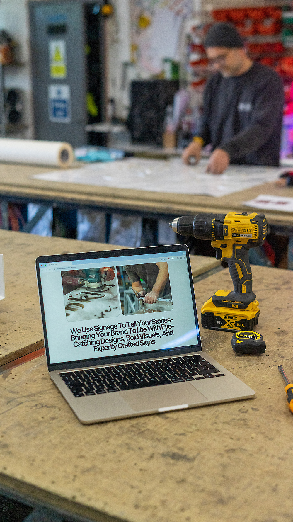 Signage website redesign - photo showing an open Macbook air on a workbench alongside a tape measure and power drill. The laptop has the Ellis Signs about page open on the screen.